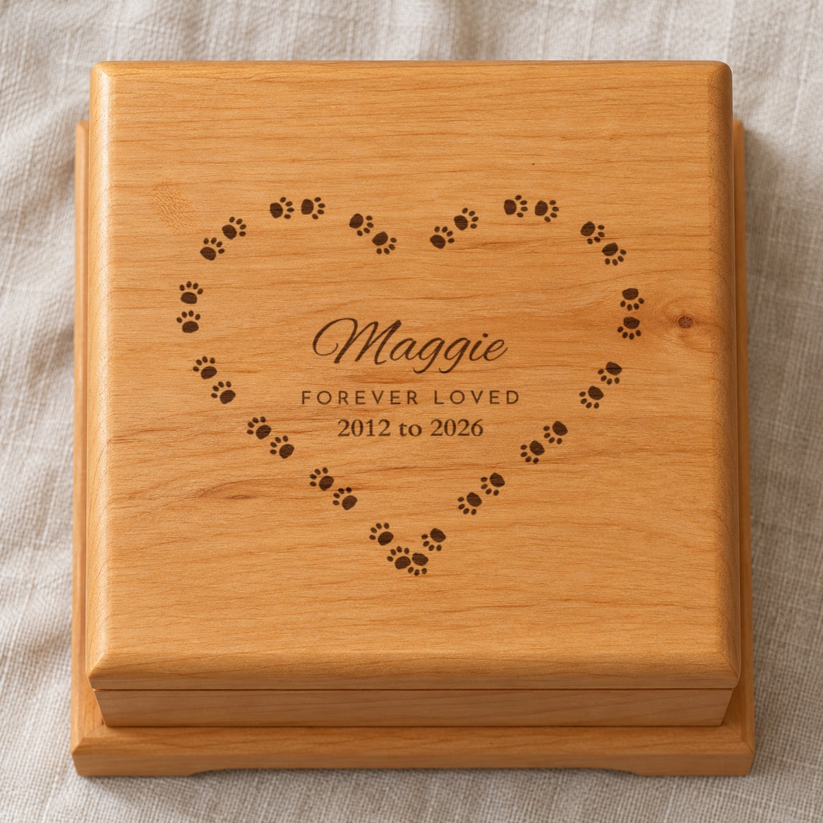 Lid of a personalized pet urn seen from above: Maggie, Forever Loved and 2012 to 2026 laser engraved inside a heart of paw prints on solid red alder