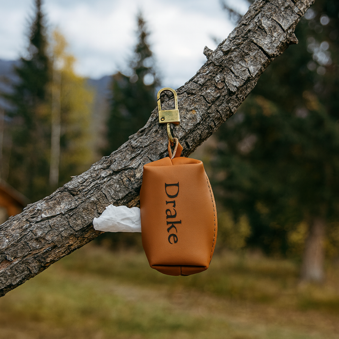 Tan leather waste bag holder with sample name "Drake" engraved, attached to tree branch with brass clip and containing white waste bags
