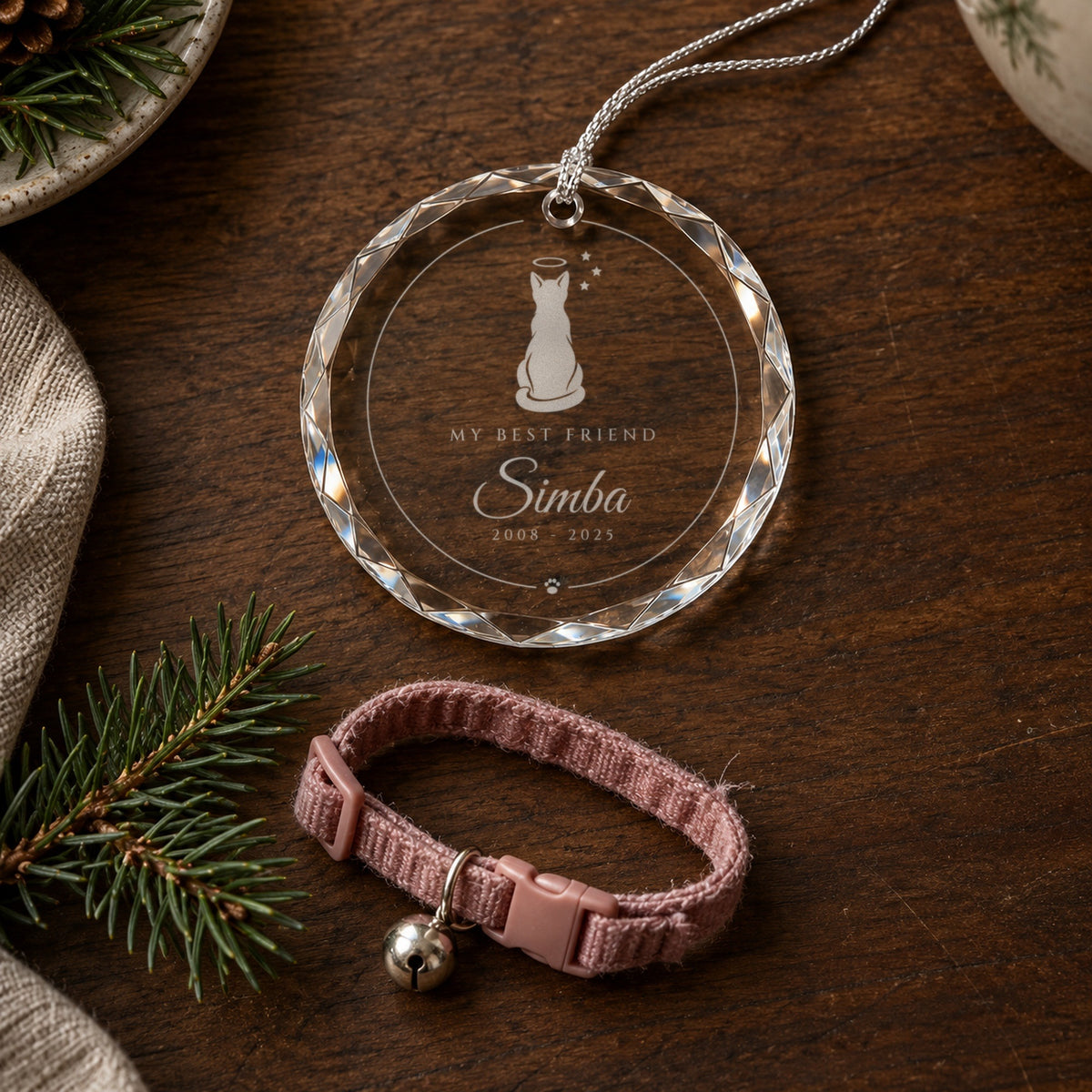 The engraved cat memorial ornament lying beside a little collar with a bell and a pine sprig on a walnut table, reading My Best Friend, Simba