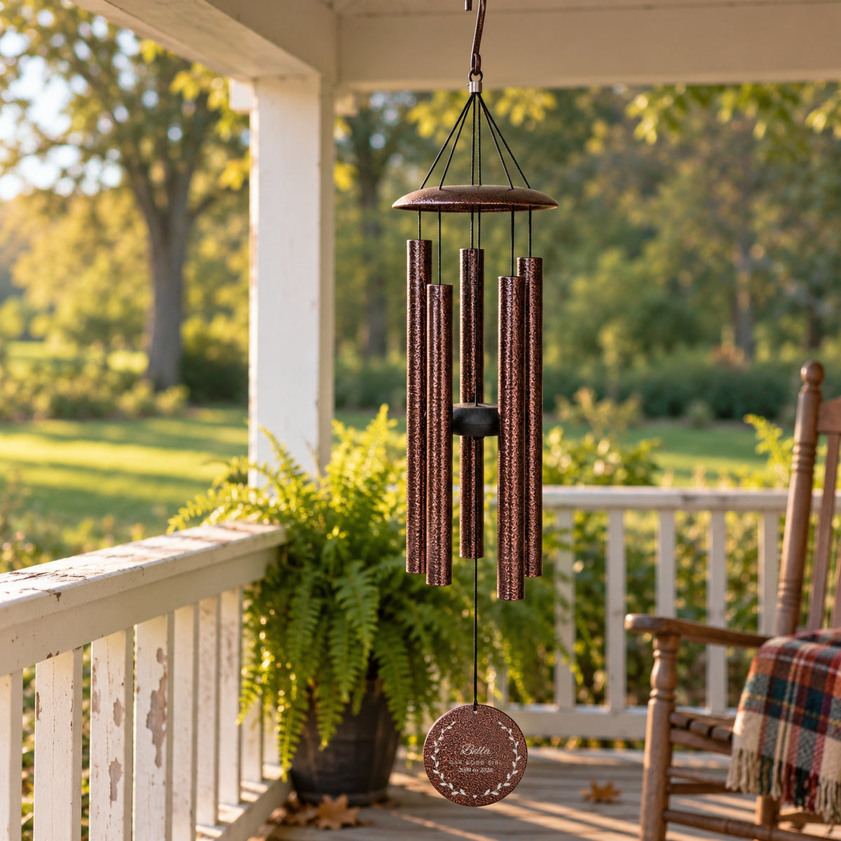 Copper pet memorial wind chime hanging on a front porch beside a fern and a rocking chair, engraved with Bella, Our Good Girl and 2009 to 2026 in a leaf wreath