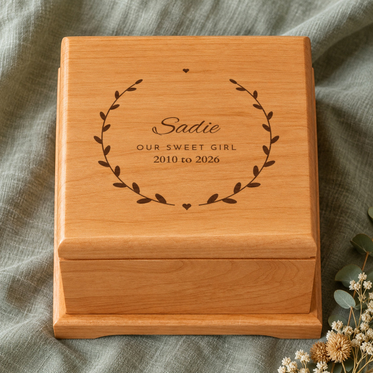 Close-up of a pet urn lid engraved with Sadie, Our Sweet Girl and 2010 to 2026 inside a leaf wreath with two small hearts, on sage linen with dried flowers