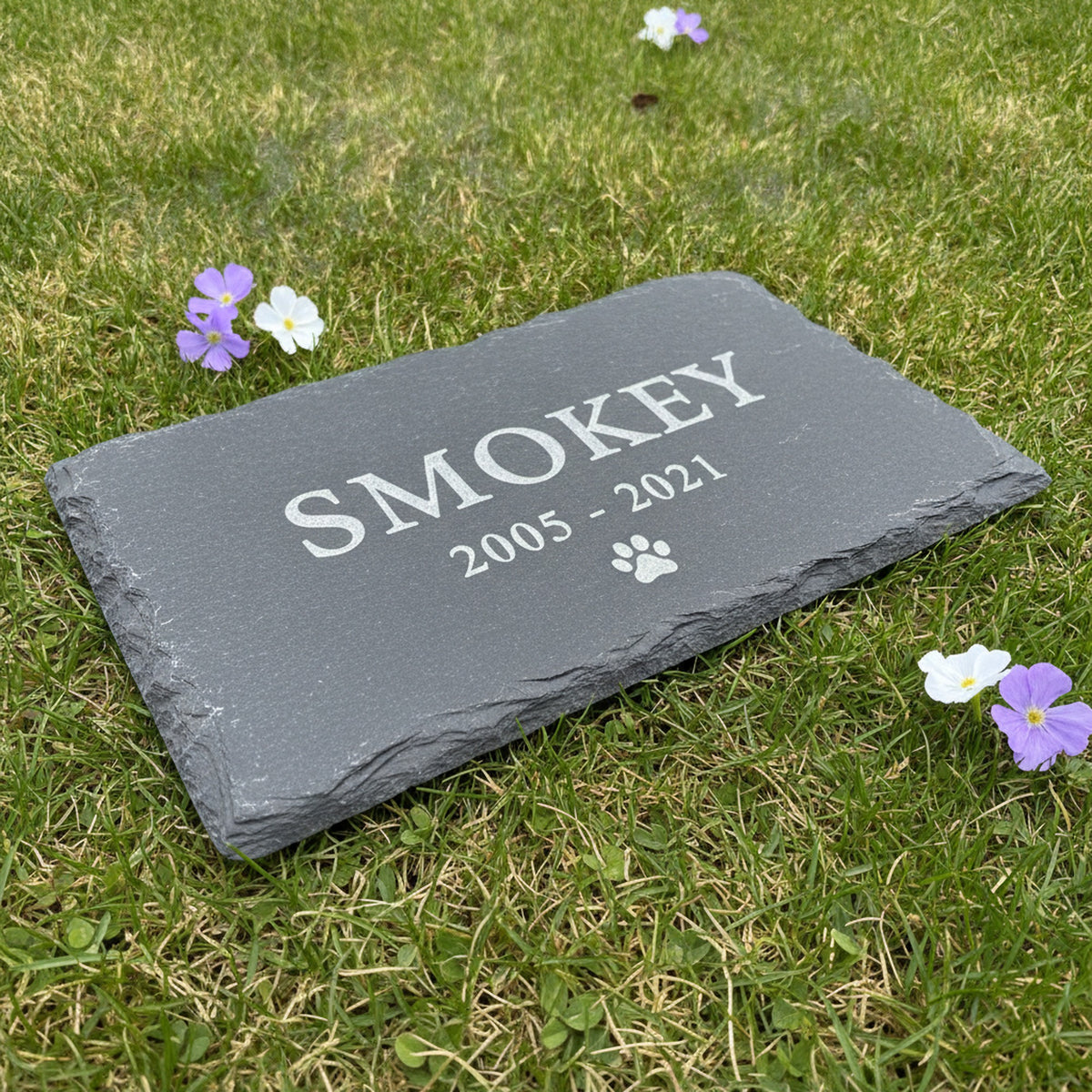 Custom slate pet memorial marker lying flat on garden grass among small purple and white flowers, engraved with a pet name, years and a paw print