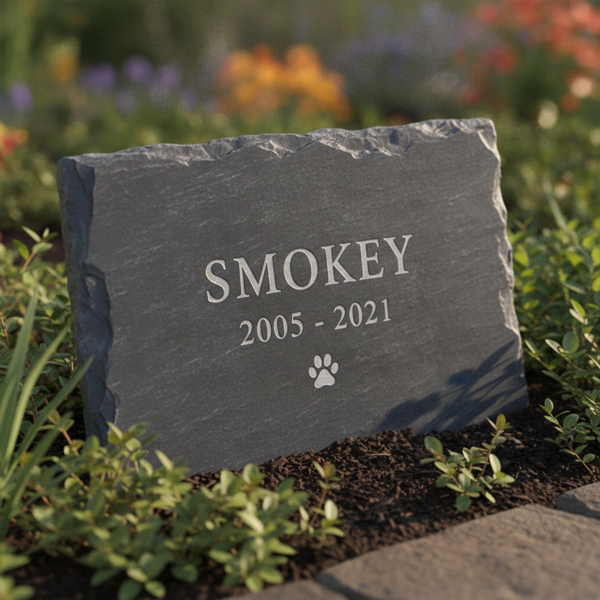 Personalized slate pet grave marker standing in a garden bed, laser engraved with the sample name Smokey, the years 2005 to 2021 and a small paw print