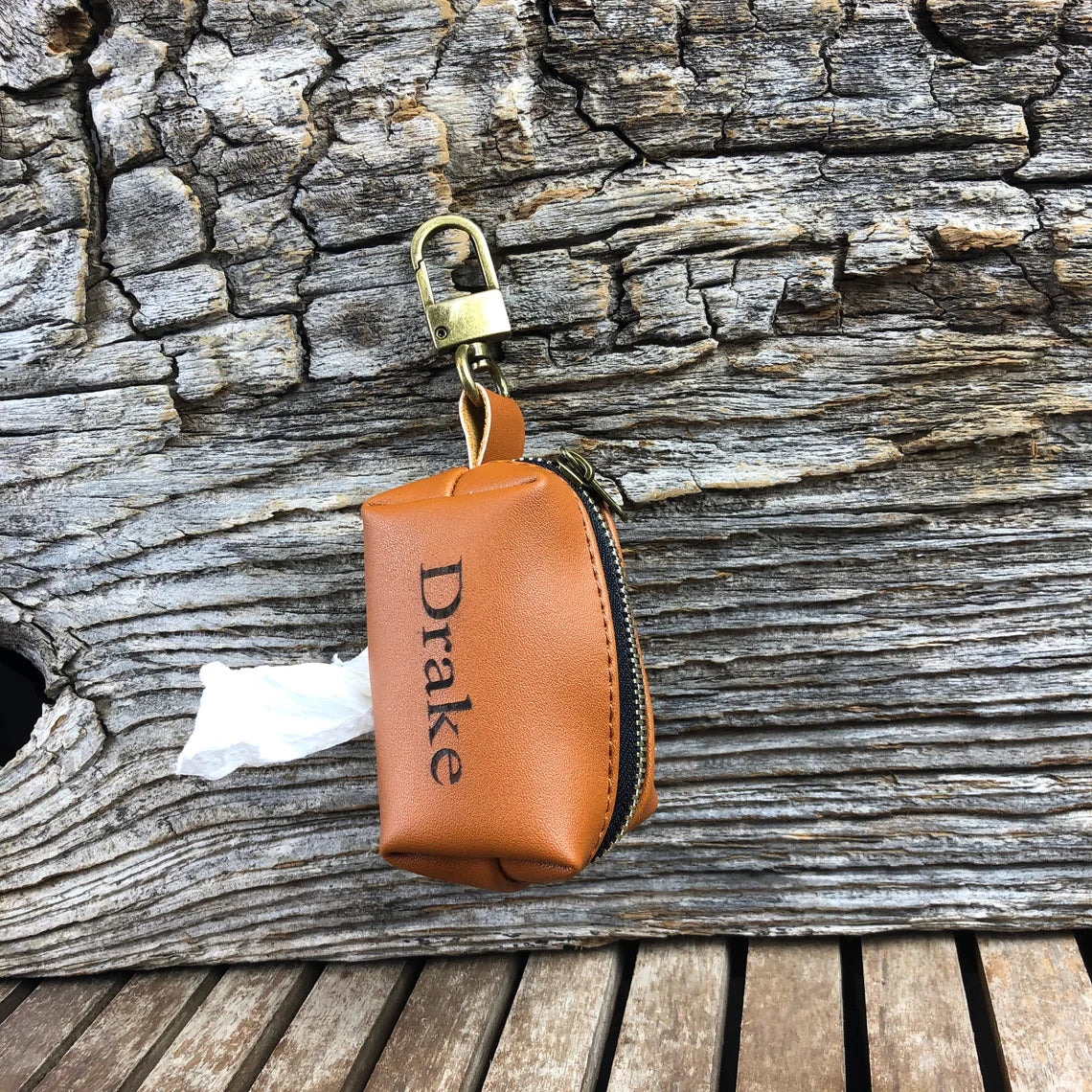 Tan leather waste bag holder with sample "Drake" engraving, brass clasp, and attached bag, displayed on weathered wood texture.