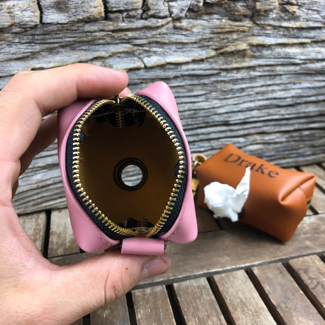 Pink leather dog waste bag holder with gold zipper, held open in hand to show interior, displayed on weathered wood surface with tan leather holder in background