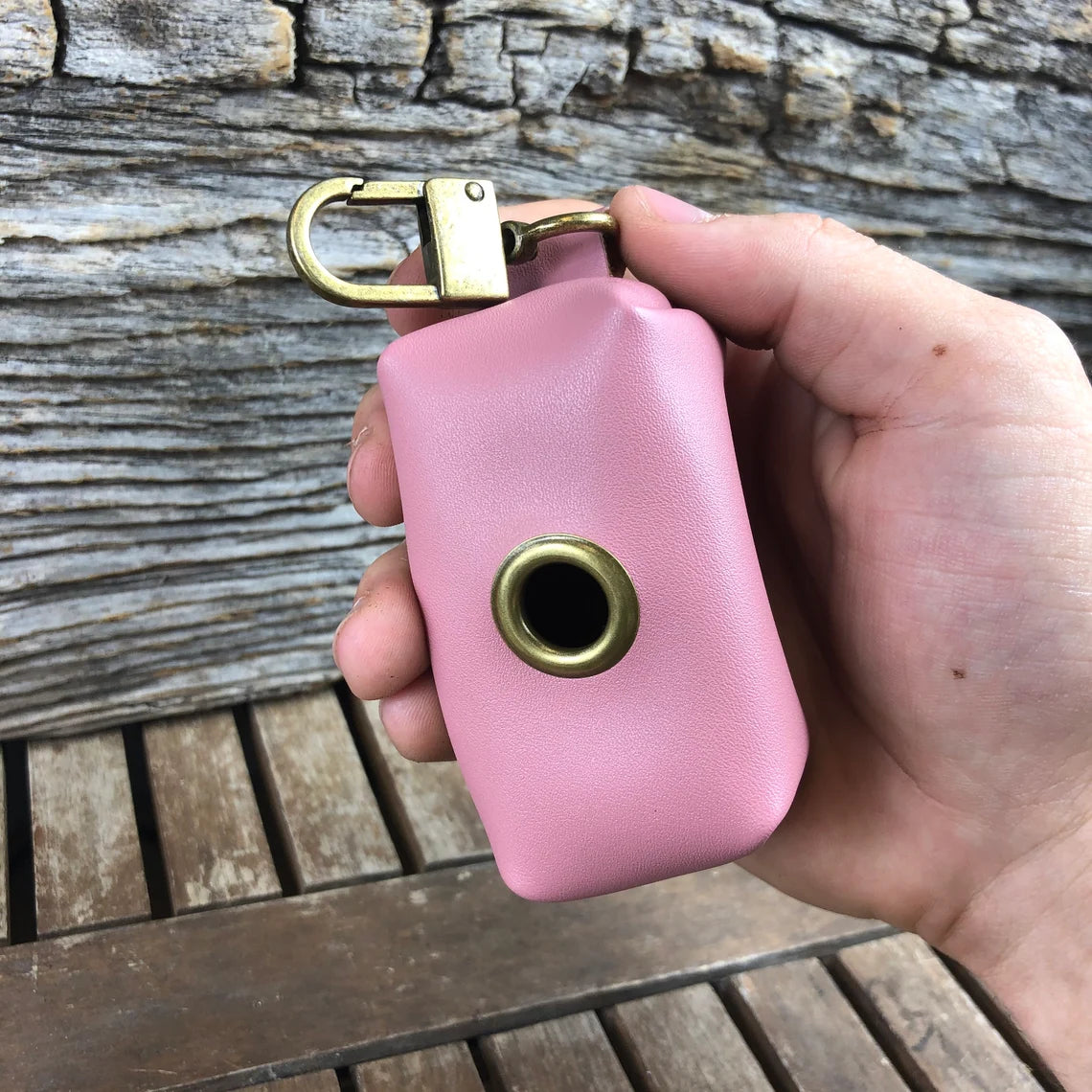 Pink leather waste bag holder with brass carabiner clip and eyelet, held in hand against weathered wood background