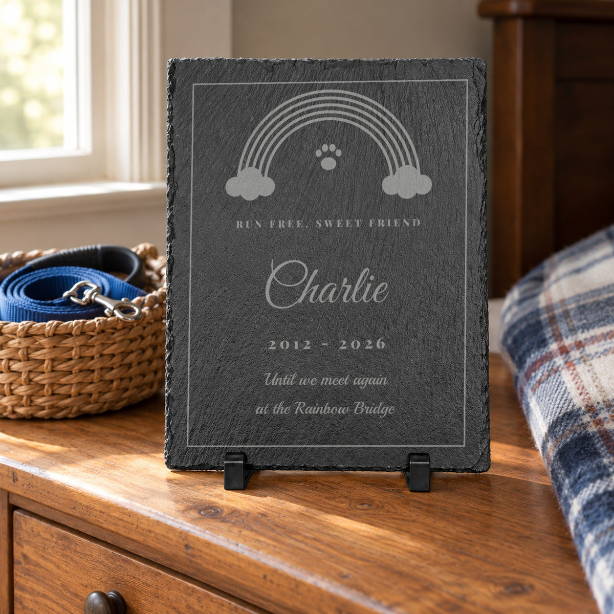 Personalized rainbow bridge pet memorial engraved Charlie 2012 - 2026 on a pine dresser beside a basket with a blue dog leash and a plaid blanket