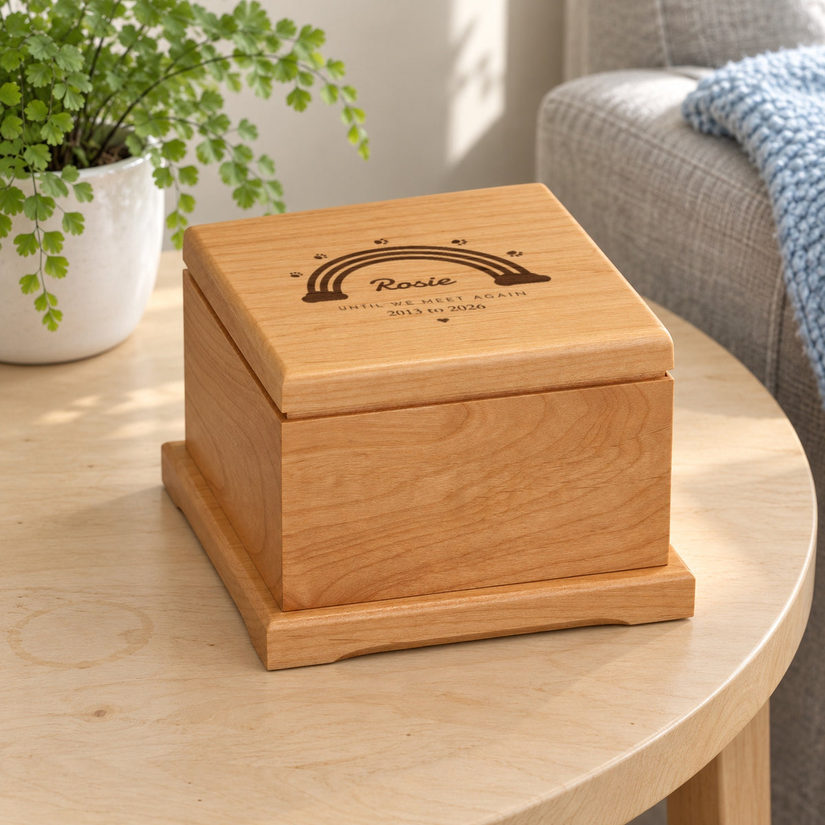 Personalized rainbow bridge pet urn on a birch side table beside a fern, the wood lid engraved with Rosie, Until We Meet Again and 2013 to 2026 under a rainbow