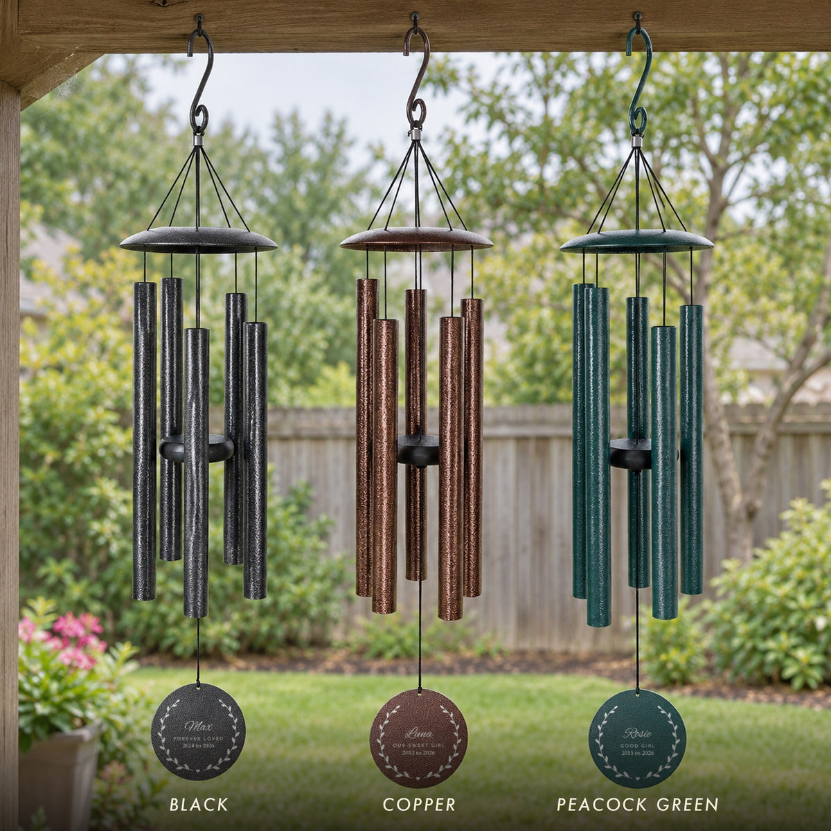 Three pet memorial wind chimes in black, copper and peacock green on a porch, engraved with the names Max, Luna and Rosie inside a leaf wreath