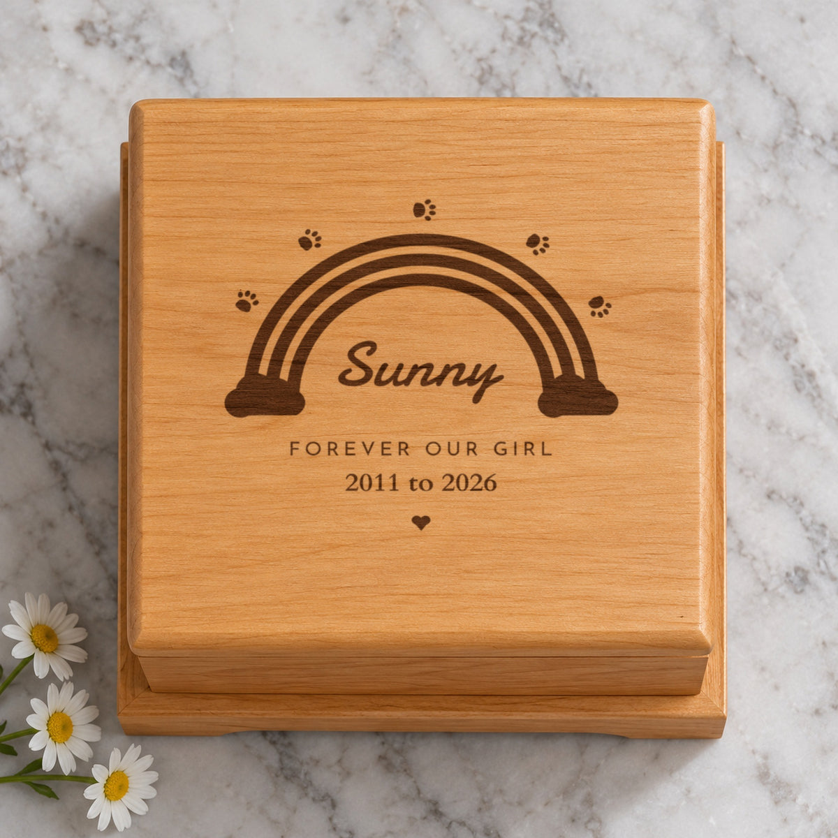 Close-up of a pet urn lid engraved with a rainbow, paw prints and a heart, with Sunny, Forever Our Girl and 2011 to 2026, on a marble counter with daisies