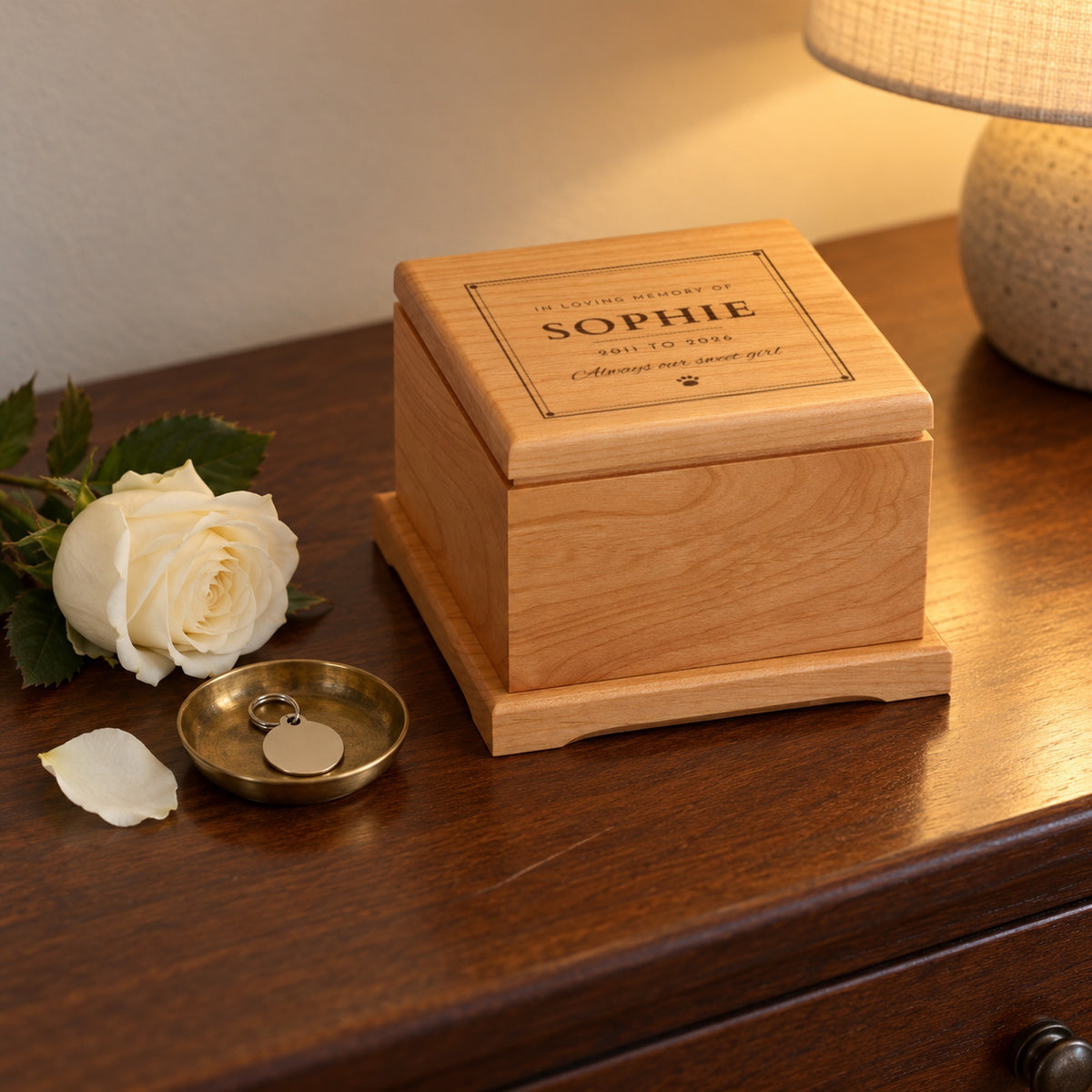 Wooden remembrance pet urn on a walnut dresser beside a white rose and a brass dish with a pet ID tag, the lid engraved with Sophie and Always our sweet girl